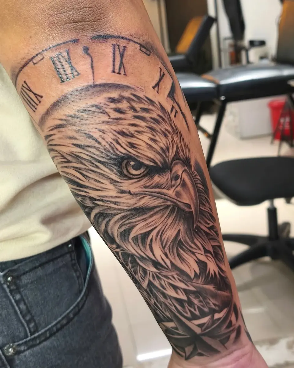 Tatuaje en realismo de un águila con un reloj de números romanos en el antebrazo, hecho por Jenn Tatts en Cali