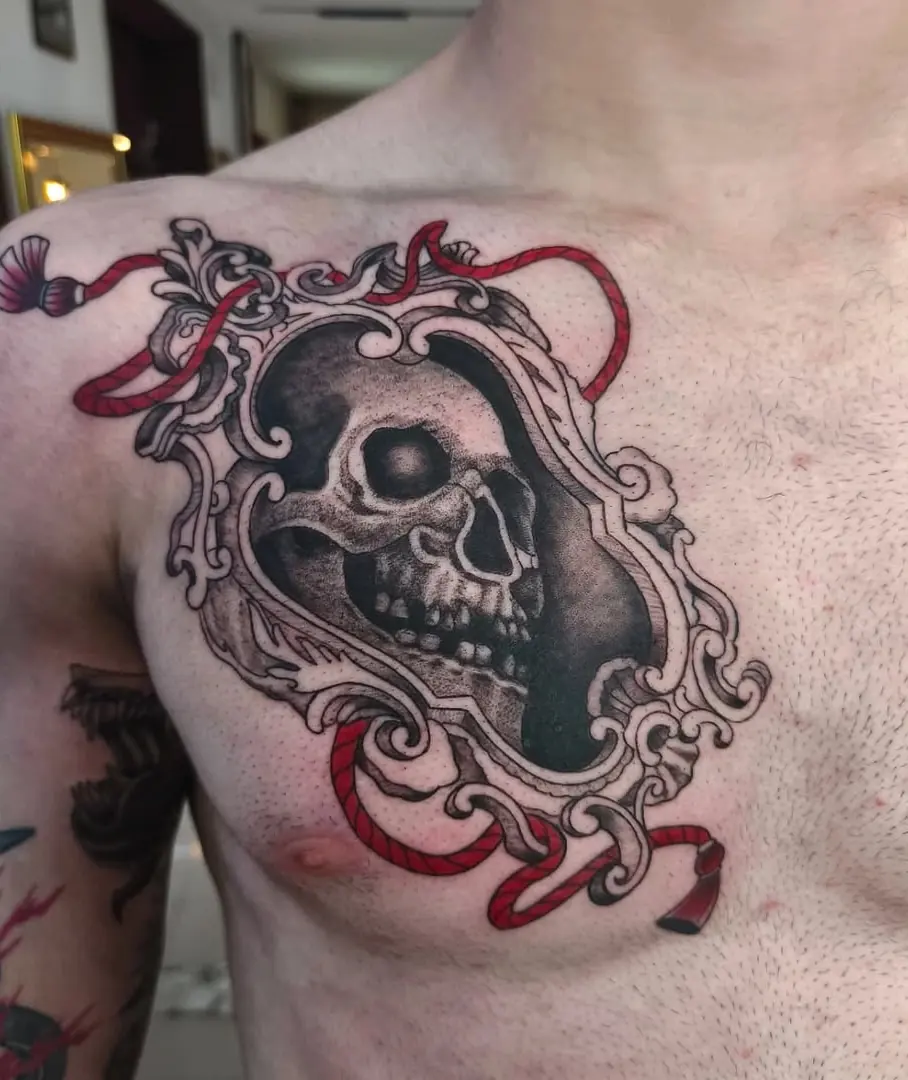Tatuaje de una calavera dentro de un marco barroco con cintas rojas en el pecho, por Jenn Tatts