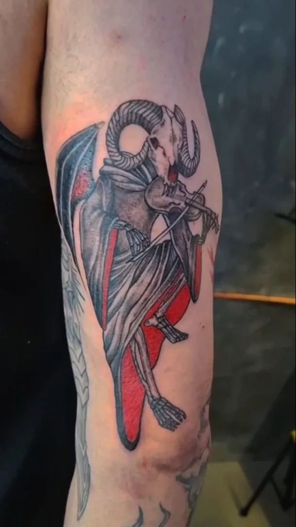 Tatuaje en black and grey de una figura con cuernos de carnero y una capa roja, en el brazo, por Jenn Tatts