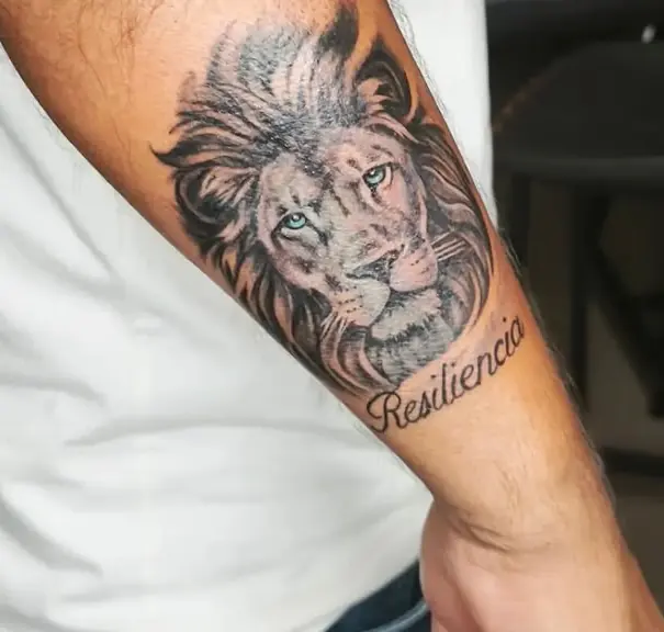 Tatuaje en realismo de un león con la palabra Resiliencia en el antebrazo, por Jenn Tatts