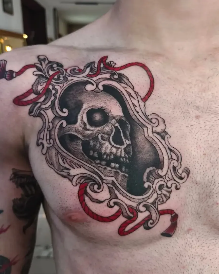 Tatuaje de una calavera dentro de un marco barroco con cintas rojas en el pecho