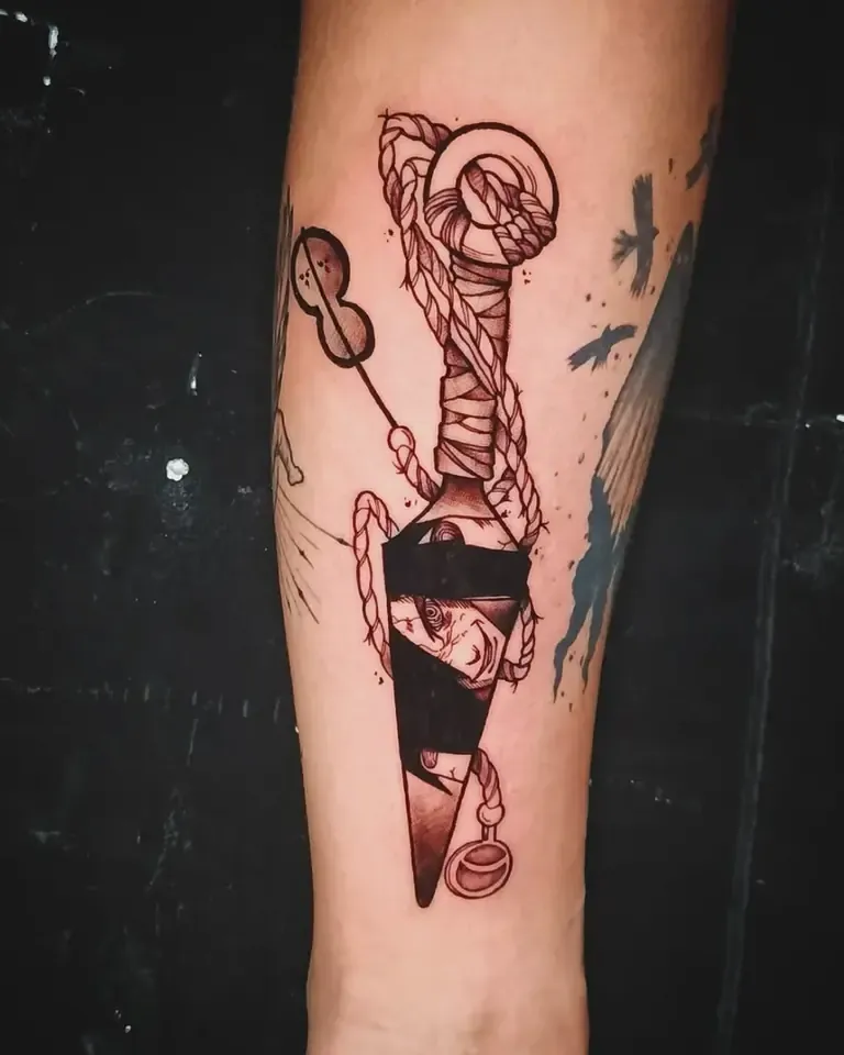Tatuaje de estilo tradicional con una daga envuelta en cuerda y detalles en rojo, en el antebrazo