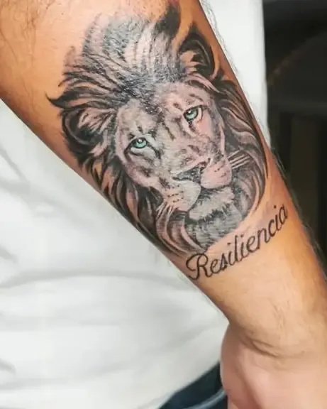 Tatuaje en realismo de un león con la palabra Resiliencia en el antebrazo